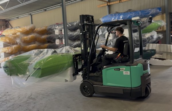 En mann kjører truck inne på et lager og frakter en grønn båt pakket inn i plast. I bakgrunnen ligger båter i flere farger stablet langs veggene.