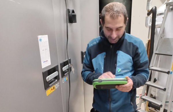 Mannen står foran ventilasjonsanlegget og ser på måleapparatet han holder. Han har arbeidsklær og blå joggesko, og står i et teknisk rom med rør og utstyr i bakgrunnen.