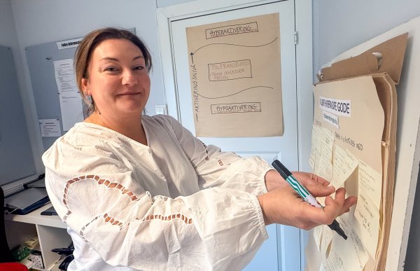 Kvinne i hvit bluse står innendørs og skriver med tusj på et stort ark som henger på veggen. Flere ark med håndskrevne notater henger rundt henne.