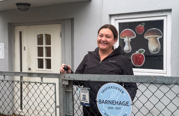 Smilende kvinne med mørkt hår og svart jakke står bak en metallport. På porten henger et blått, rundt skilt, og bak henne er et vindu dekorert med sopper og epler.
