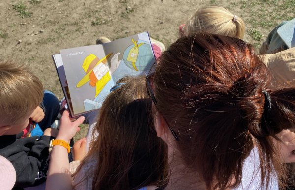 En voksen sitter sammen med flere barn ute og holder opp en bildebok. Barna sitter tett rundt og ser på illustrasjonene mens hun leser.