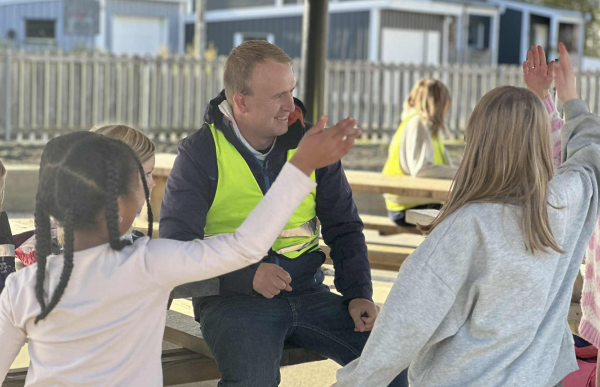 Barne- og ungdomsarbeider Einar Johansen i gul vest, prater med barna i friminuttet. Han sitter på en benk mens barna står. En voksen person med gul vest sitter ved et piknikbord sammen med flere barn. Barna har hendene løftet, og de ser ut til å være i samtale eller aktivitet