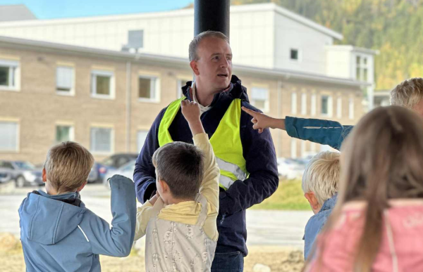 Barne- og ungdomsarbeider Einar Johansen i gul vest står og prater med barna i friminuttet. Mann med gul vest står under et overbygg med flere barn rundt seg. Barna rekker opp hendene, og bord er synlige i forgrunnen.
