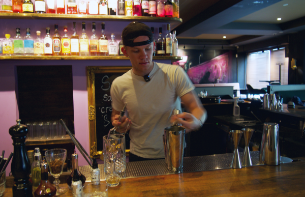 En ung mann med svart caps og hvit t-skjorte står bak en bar med flasker i bakgrunnen. Foran han står en shaker i sølv. Han er i gang med å helle noe over i shakeren. 