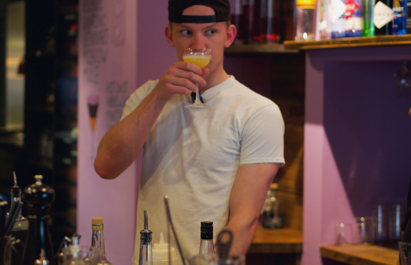 En ung mann med svart caps og hvit t-skjorte står og lukter på et cocktailglass med gult innhold. Rundt han står ulike flasker og glass