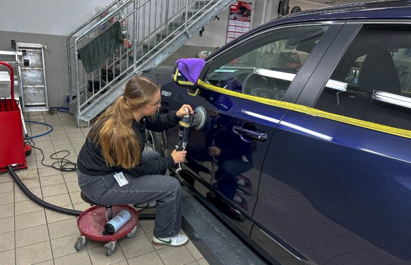 Dame med grå bukse og svart genser, og brunt hår i hestehale, sitter på en krakk og bruker poleringsmaskin på en blå bil.