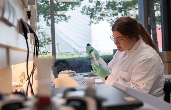Dame med hvit frakk sitter ved en benk på et laboratorium. Hun har på seg grønne plastikkhansker og bruker en pipette.