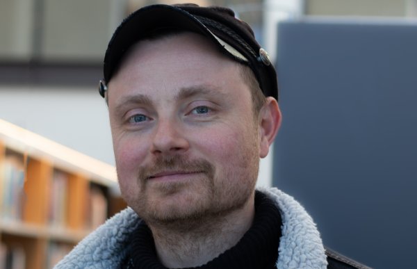 En mann i svart genser og svart jakke med hvitt for. Han har en svart caps på hodet. Han er inne.