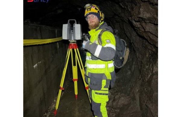 Mann i full refleks- og verneutstyr, med hjelm, briller, jakke, bukse og vernesko, står i det som ser ut som en tunnel eller gruve. Ved siden av ham står måleutstyr, og han poserer med tommelen opp.