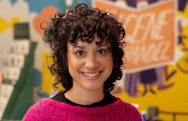 Ei dame med svart og krøllete hår. Hun har rosa genser på. Hun smiler og ser rett i kamera. Hun er i en bokhandel.