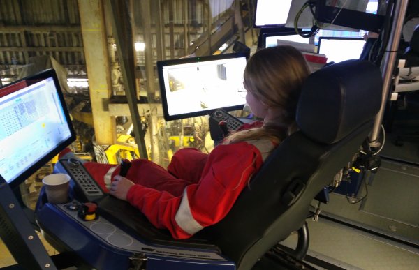 En kvinne i rød arbeidsdress sitter i en stol foran to dataskjermer 