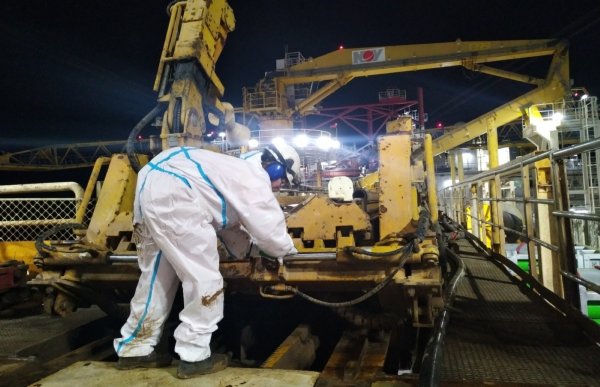 En kvinne med hvit kjeledress, hjelm, hørselvern og vernebriller bøyer seg ned og smører en stor maskin