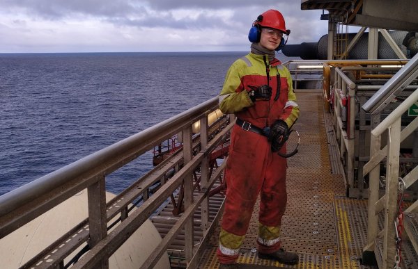 En mann står på boredekk med hjelm og orange arbeidsdress med havet i bakgrunn