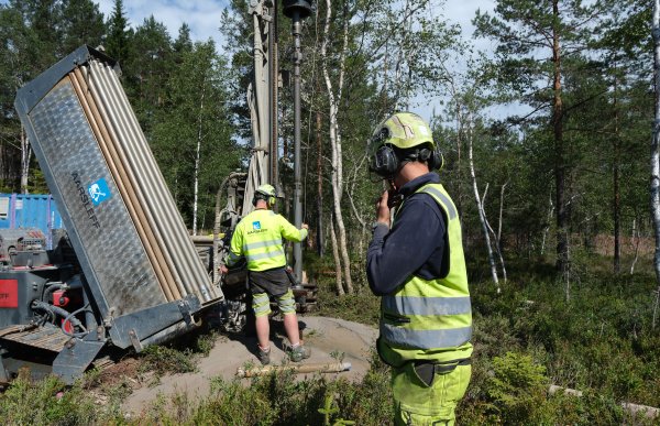 To brønnborere arbeider ved en borerigg i et skogsområde. Den ene betjener riggen, mens den andre følger med iført hjelm, hørselvern og refleksklær. Bildet viser brønnboring i felt.