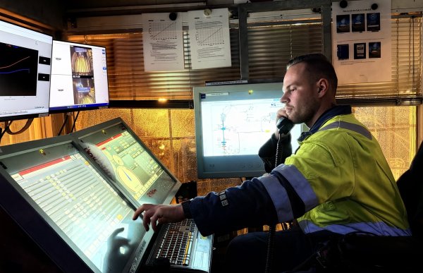 En mann i gul arbeidsdress sitter ved flere dataskjermer og snakker i telefonen