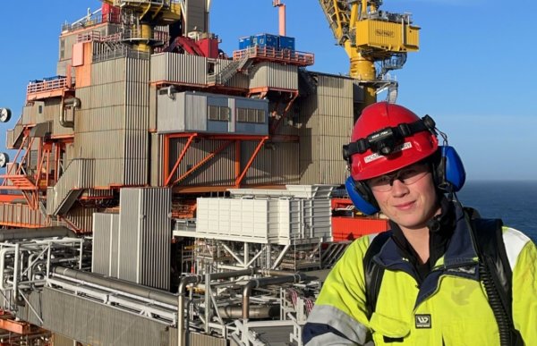 En mann i gul arbeidsdress, rød hjelm og øreklokker står på en borerigg med havet i bakgrunnen
