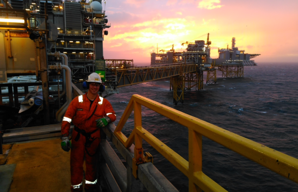 En mann i orange arbeidsdress og hjelm står på et dekk med havet og borerigg bak