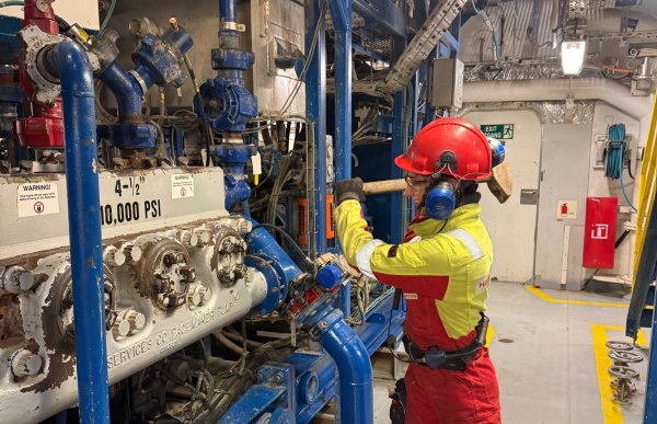 En kvinnelig offshorearbeider i vernehjelm og hørselsvern bruker en stor hammer på blått høytrykksutstyr inne i et teknisk rom på en offshoreinstallasjon. Rundt henne står rør, ventiler og tungt industrielt utstyr.