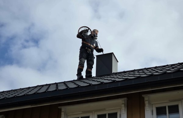 En person i svart arbeidsdress og maske i ansiktet står ved ei murpipe på taket av et hus og sender en wire ned i murpipa.