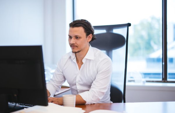 En mann i hvit skjorte sitter ved et skrivebord foran en dataskjerm. Han ser konsentrert ut mens han jobber på kontor, med store vinduer og dagslys i bakgrunnen.