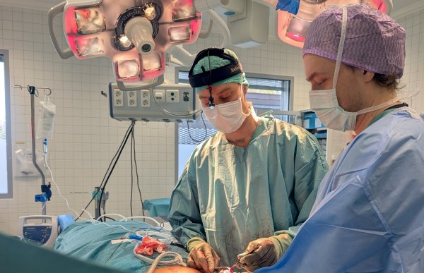 To personer med munnbind og hette på hodet holder et instrument inntil en kropp som ligger på en operasjonsbenk