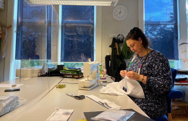 En dame sitter ved et bord og syr på en hvit skjorte. På bordet står en symaskin og andre sy-verktøy. 