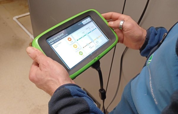 Et nærbilde av mannens hender som holder et grønt måleapparat med skjerm. På skjermen vises et teknisk diagram over ventilasjonsanlegget.