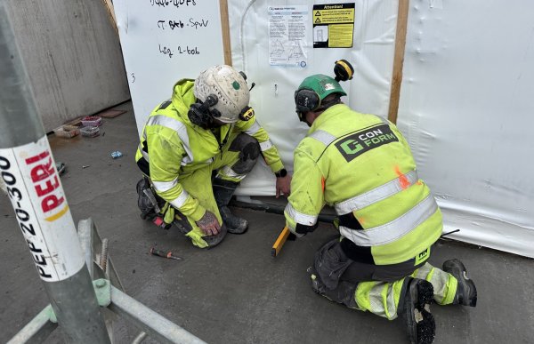 To byggarbeidere i gule refleksklær fra firmaet «Con Form» sitter på kne og arbeider foran en stor, hvit innpakket prefabmodul merket «Bano Prefab». Den ene har hvit hjelm, den andre grønn. De måler eller justerer noe langs kanten av modulen. Rundt dem ligger verktøy og byggematerialer på betonggulvet.
