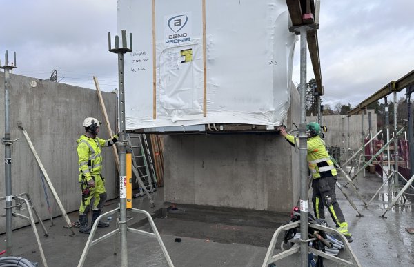 To byggarbeidere i gule refleksklær fra «Con Form» monterer en stor, hvit modul. Modulen henger i en kran og senkes på plass mellom betongvegger. Den ene arbeideren med hvit hjelm står til venstre og styrer plasseringen, mens den andre med grønn hjelm holder i modulen fra høyre side. Det er grå himmel og våt betong på bakken.