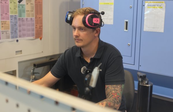 En mann sitter ved en dataskjerm i et industriverksted. Han har et rosa og svart hørselsvern på hodet og bruker en mørk arbeids­pique. I bakgrunnen står store CNC-maskiner og oppslag med tekniske tabeller. Personen jobber konsentrert foran skjermen.