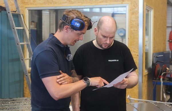 To personer står i et verksted og ser sammen på et utskrevet ark. Den ene peker på dokumentet mens den andre holder det. Begge bruker arbeidsklær, og den ene har hørselsvern på hodet. Rundt dem sees metallmaskiner, verktøy, en stige og vinduer inn til et annet rom.