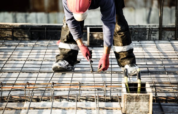 En person iført rød hjelm, hansker og øvrig arbeidstøy bøyer seg ned og jobber med armeringsjern på en byggeplass.