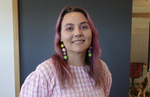 Jente med rosa hår og rosa og hvit rutete kjole som smiler foran en vegg.