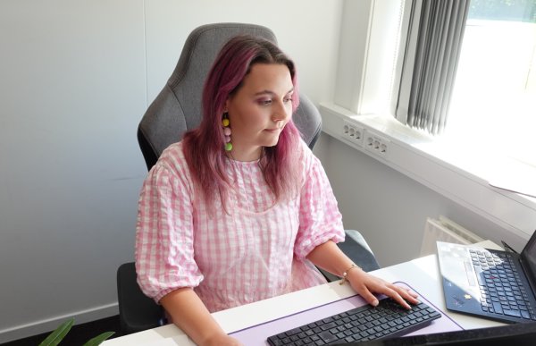 Jente med rosa hår og rosa og hvit rutete kjole sitter foran en pc-skjerm og jobber.