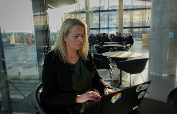 Kvinne sitter på stol med svart dressjakke og en pc foran seg. I bakgrunn sitter det mennesker samlet rundt et bord. 