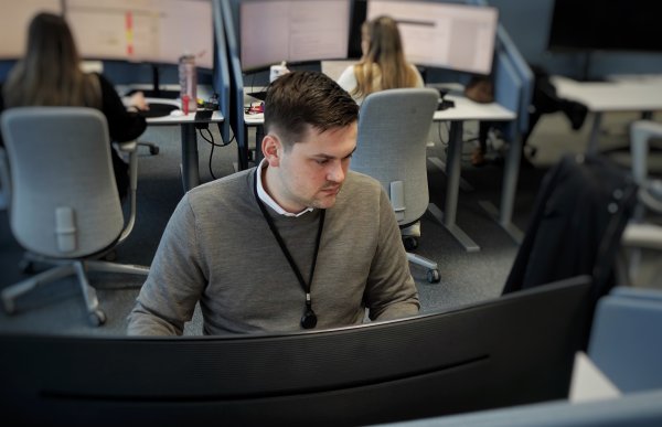 Mann i grå genser og brunt hår sitter foran en skjerm, to jenter sitter i bakgrunnen og jobber.