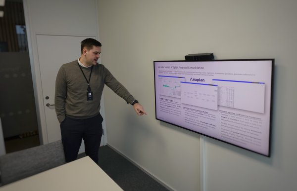 Mann i grå genser og svart bukse står oppreist og peker mot en skjerm i et møterom