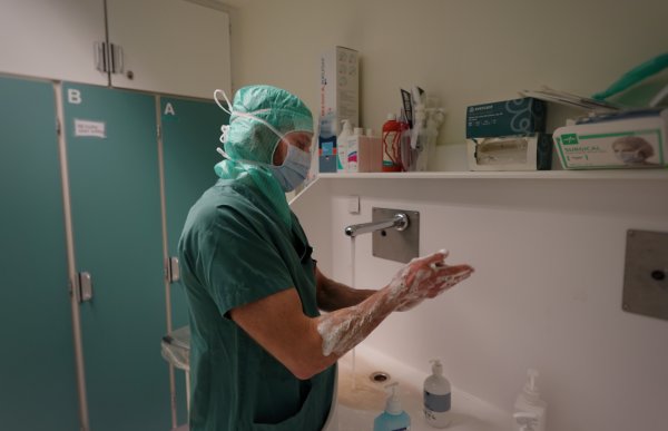 Mann i grønn arbeidsuniform med grønn hette og munnbind. Han vasker hendene. 