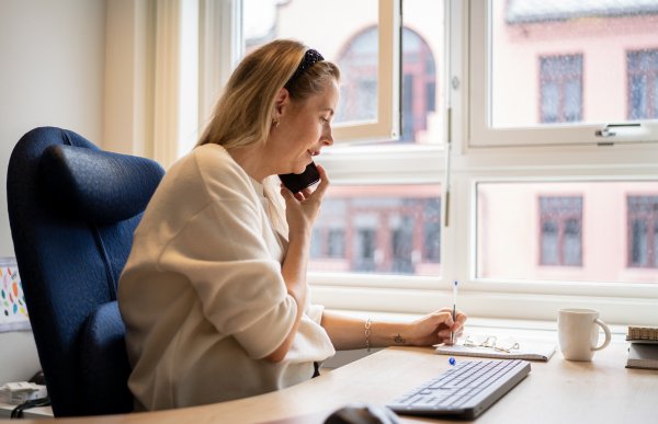 Kvinne i hvit genser ved et skrivebord snakker i telefon.