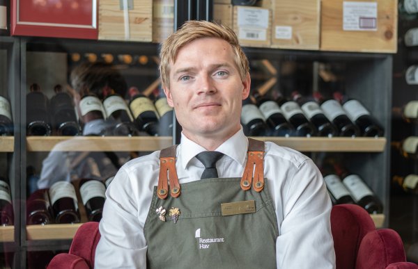 Mann i hvit skjorte og med et grønt kelnerforkle sitter foran et vinskap.