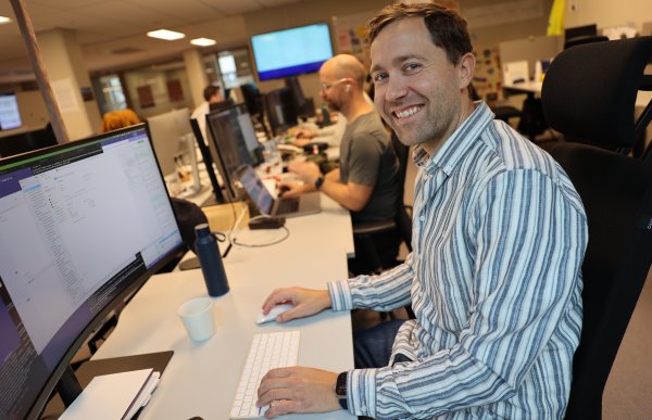 En mann sitter foran en dataskjerm på et åpent kontor. Han smiler mot kameraet mens han holder hånden på tastaturet. Rundt ham sitter kolleger som jobber ved sine stasjoner.