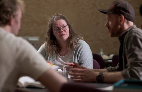 En kvinne med briller sitter rundt et bord og har en samtale med to andre mennesker. De har på seg vanlige klær og ser fokuserte ut. På bordet er det noe papirer og en person sitter med en penn i hånda.