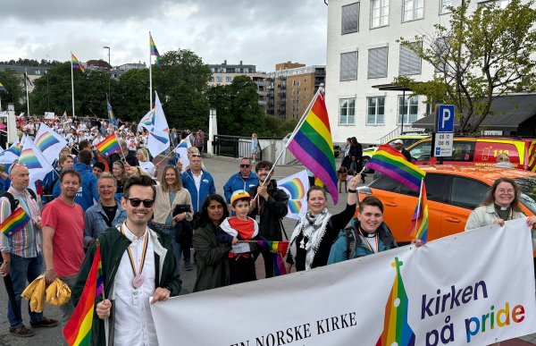 Mange mennesker står i en pride-parade. Folk smiler og holder pride-flagget. Menneskene som står foran holder en banner som sier "kirken på pride"