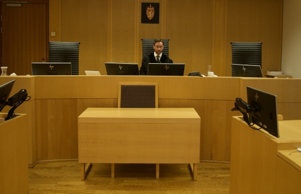 En mann i dommerkappe sitter på en stol i en rettssal. Stolen er plassert i midten av to andre, på en forhøyning i rommet. Rommet er kledd i tre, og har diverse skjermer på bordene. Bak mannen henger det norske riksvåpenet på veggen.