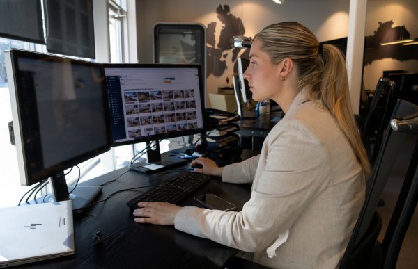 Ei dame med lyst og langt hår. Hun har på en beige jakke. Hun sitter foran to pc-skjermer og ser på dem. Hun er på et kontor.