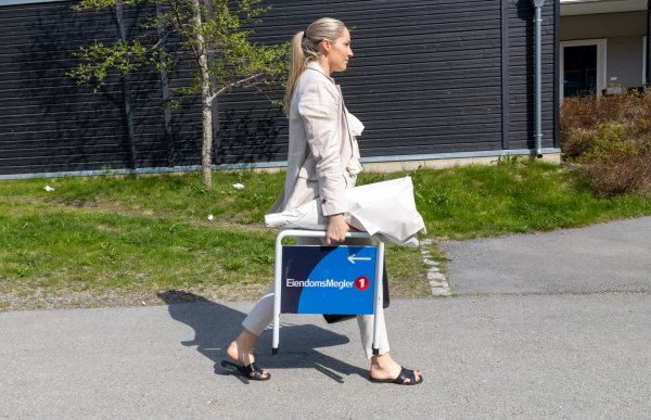 Ei dame med lyst og langt hår. Hun har på en beige jakke og en beige bukse. Hun bærer blomster pakket i papir og et skilt med teksten "EiendomsMegler 1". Hun går på et fortau. I bakgrunnen er en svart bygning.