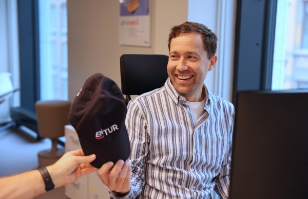 En mann sitter ved skrivebordet sitt og smiler mot en kollega som rekker ham en mørk caps med logoen «Entur». Lyset fra vinduet faller inn over pulten og gir scenen et varmt preg.