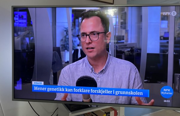 En mann med briller og kort, mørkt hår sitter i et studio og snakker i en mikrofon. Han har på seg en lyseblå skjorte med ståkrage. Nederst i bildet vises NRK-logo, klokkeslett og teksten «Mener genetikk kan forklare forskjeller i grunnskolen.