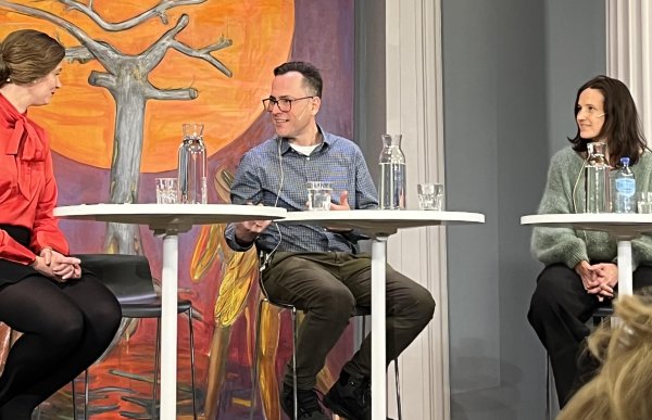Tre personer sitter ved et rundt bord på en scene. Mannen i midten har briller, rutete skjorte og mikrofon i øret. I bakgrunnen henger et stort maleri av en fugl foran en rød sol og et tre. På bordet står vannkarafler og glass.