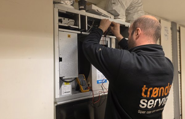 En mann i svart arbeidsjakke holder et måleinstrument inni en varmepumpe på en vegg.